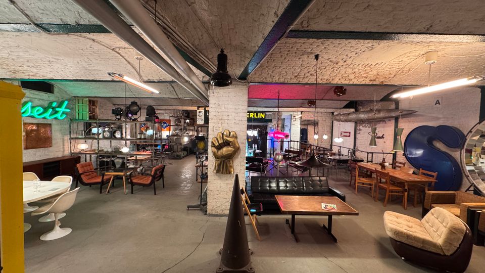 Industrieller Innenraum mit gemauerten weißen Wänden, sichtbaren Rohren und einer Kombination aus Retro- und modernen Möbeln, darunter Ess- und Loungetische sowie Sessel. Im Hintergrund befinden sich Neon-Schilder, Regale mit beleuchteten Objekten und dekorative Elemente wie eine große, blaue Skulptur in Form eines „S“.