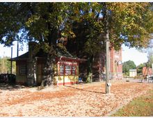 Bahnhofsumgebung mit herbstlich bedecktem Boden, einem kleinen gelb-roten Gebäude mit der Aufschrift „Erfrischungs-Halle“ und einem größeren Backsteingebäude im Hintergrund. Große Bäume spenden Schatten, und rechts ist eine Uhr sowie etwas Straßenmobiliar sichtbar.