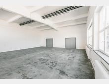 Ein leerer, heller Raum mit weiß gestrichenen Wänden, grauem Betonboden und großen Fenstern auf der rechten Seite, die Tageslicht hereinlassen. An der Stirnwand befinden sich zwei graue Metalltüren, und die Decke zeigt freiliegende Träger.