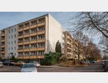 Mehrstöckiges Wohngebäude mit gelben Balkonen in einer Wohnsiedlung. Im Vordergrund geparkte Autos, Gehwege und spärlich belaubte Bäume entlang der Straße.