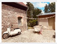 Gepflasterter Innenhof zwischen einem Backsteingebäude mit rotem Ziegeldach und einem Holzgebäude mit Doppeltür. Im Hof stehen ein abgedeckter Tisch mit roten Stühlen sowie ein weißes Moped; im Hintergrund sind Bäume und blauer Himmel zu sehen.