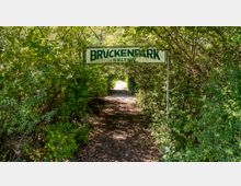 Eingang zu einem Waldweg im Brückenpark Geltow, mit einem grünen Schild, das den Parknamen anzeigt, umgeben von dichtem Grün und Bäumen. Der Weg ist mit Blättern bedeckt und führt durch eine natürliche Umgebung.