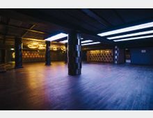 Großer, leerer Innenraum mit Holzboden, großen Säulen und einer modernen Decke mit leuchtenden rechteckigen Lichtpaneelen. An den Wänden sind geometrische Muster und Dekorationen erkennbar, die mit warmem Licht beleuchtet werden.