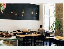 Ein gemütliches Café mit Holztischen und schwarzen Stühlen, einer schwarzen Wand mit hängenden Glühbirnen als Beleuchtung, einem großen Fenster mit Tageslicht und einer sichtbaren Backsteinmauer. Im Vordergrund stehen Tische, und im Hintergrund sitzt eine Person an einem Laptop.