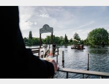 Blick auf eine Holzplattform am Wasser mit dem Schild „Freiheit Fünfzehn“. Im Hintergrund ein kleines Boot auf einem Fluss, umgeben von Bäumen unter einem klaren Himmel.