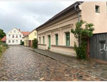 Kopfsteinpflasterstraße mit historischen Wohnhäusern, darunter ein beiges Gebäude mit grünen Fensterrahmen und dekorativen Fensterstürzen. Im Hintergrund weitere Häuser mit roten Ziegeldächern und vereinzelten Bäumen.
