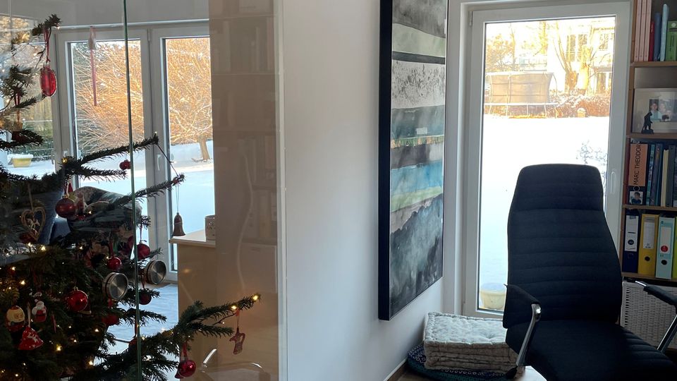Heller Innenraum mit Schiebetür aus Glas, Holzoptik-Boden und Blick durch große Fenster und eine Glastür in einen verschneiten Garten. Rechts stehen ein schwarzer Bürostuhl auf einem Teppich und ein Regal, links ist ein geschmückter Weihnachtsbaum zu sehen.