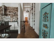 Helle Wohnung mit offener Küche und Flur. Die Küche hat eine Backsteinwand, moderne Küchenschränke und eine Edelstahl-Dunstabzugshaube; im Flur stehen ein Fahrrad und Regale mit Büchern, während eine verzierte, türkisfarbene Tür im Vordergrund sichtbar ist.