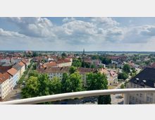 Blick auf eine Kleinstadt mit roten Ziegeldächern und mehrstöckigen Wohnhäusern, umgeben von Bäumen und Grünflächen. Im Hintergrund sind eine Kirche mit spitzem Turm und flache Felder unter einem bewölkten Himmel zu sehen.