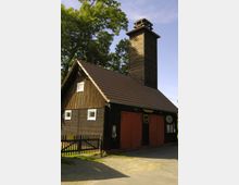 Holzgebäude mit einem rot gedeckten Satteldach, zwei großen roten Toren und kleinen weißen Fenstern, daneben ein hoher hölzerner Turm mit Glocken. Im Vordergrund ein kleiner Zaun und ein Telefon an der Wand, umgeben von Bäumen und einer sonnigen Umgebung.