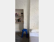 Minimalistischer Innenraum mit weiß gestrichener Backsteinwand und einem eingebauten Regal mit Büchern und kleiner Tischlampe. Daneben stehen ein blauer Hocker auf Betonboden und ein Bett mit weißer Bettwäsche am rechten Bildrand.