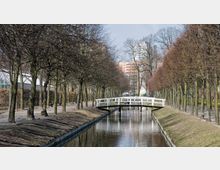 Parklandschaft mit einem geraden Wasserkanal, der von kahlen Bäumen gesäumt ist. Über den Kanal führt eine schmale, weiße Holzbrücke, im Hintergrund sind Gebäude und ein Gewächshaus sichtbar.