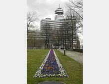 Mehrstöckiges Bürogebäude mit einer markanten, gestuften Architektur und einer Antenne auf dem Dach, im Vordergrund eine Grünfläche mit einem Blumenbeet in Weiß, Blau und Rot, flankiert von Bäumen und einem gepflasterten Weg. Der Himmel ist bewölkt.