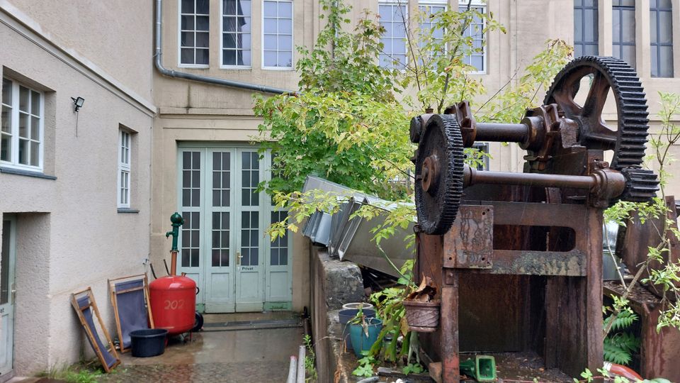Hinterhof mit alten Maschinenkomponenten, darunter ein rostiges Zahnradgerät im Vordergrund. Im Hintergrund befindet sich eine beige Gebäude-Fassade mit großen Fensterflächen und einer Tür mit der Aufschrift „Privat“, umgeben von Pflanzen und verschiedenen Gegenständen.