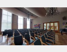 Ein moderner Veranstaltungsraum mit Reihen von gepolsterten Stühlen und hohen, bodentiefen Fenstern, die vertikale Lamellen haben. An der Decke hängen Kronleuchter, und an den Wänden befinden sich bunte Gemälde.