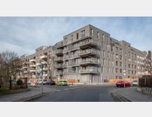 Mehrstöckiges modernes Wohngebäude mit grauer Fassade und zahlreichen Balkonen, die mit Glasgeländern ausgestattet sind. Das Gebäude steht an einer Straßenecke in einer urbanen Umgebung mit Gehwegen, parkenden Autos und einigen Bäumen.