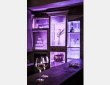 Ein stilvoller Innenraum mit beleuchteten Vitrinen, die Kristallgläser, Karaffen und Dekorationsgegenstände enthalten. Im Vordergrund stehen zwei Gläser mit Wein auf einer dunklen Holztheke.