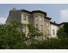 Mehrstöckiges Wohngebäude mit hellbeigen und weißen Fassaden, teilweise von Bäumen und Sträuchern im Vordergrund verdeckt. Die Architektur weist einfache dekorative Elemente und mehrere Fenster auf, vor einem klaren Himmel.