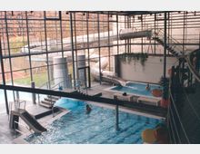 Hallenbad mit großen Glaswänden, mehreren Schwimmbecken, einer Kinderrutsche und einer langen Röhrenrutsche, die von außen ins Gebäude führt. Der Raum ist lichtdurchflutet, mit vereinzelt dekorierten Geländern und Pflanzen an den Wänden.