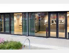 Modernes Gebäude mit großer Glasfassade, durch die ein Innenraum mit hängenden Schwarz-Weiß-Fotografien sichtbar ist. Vor dem Eingang befindet sich eine Klingel, und die Türen sind mit den Aufschriften "Eingang" und "Entrance" versehen.