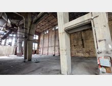 Innenansicht eines verlassenen Industriegebäudes mit sichtbaren Betonstützen, freiliegenden Stahlträgern und teils verblassten Backsteinwänden. Der Boden besteht aus unebenem Pflaster, und Sonnenlicht dringt durch hohe Fenster ein.