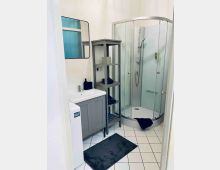 Kleines Badezimmer mit weiß gefliesten Wänden und Bodenfliesen im Rastermuster, links ein Waschbecken mit Unterschrank und Wandspiegel sowie ein schmales Regal. Rechts befindet sich eine abgerundete Glas-Duschkabine mit Handbrause; auf dem Boden liegt ein dunkler Badvorleger.