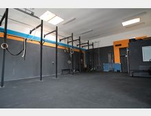 Ein moderner Trainingsraum mit dunklen Wänden und Akzenten in Blau und Orange. Es gibt Metallgerüste mit Turnringen und Kletterseilen, sowie an der Wand gelagerte Langhanteln und Fitnessgeräte.