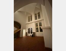 Ein heller Raum mit hohen Decken und Spitzbögen, großen Fenstern mit Sprossenteilung und verzierten Doppeltüren. Der Raum verfügt über eine Holztreppe auf der linken Seite, einen roten Fliesenboden und Heizkörper unter den Fenstern.