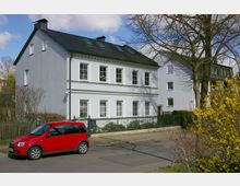 Zweistöckiges Wohnhaus mit weißer Fassade und dunklem Schieferdach, umgeben von einem schmiedeeisernen Zaun und kahlen Bäumen. Im Vordergrund steht ein rotes Auto auf der Straße, daneben Frühjahrssträucher mit gelben Blüten.