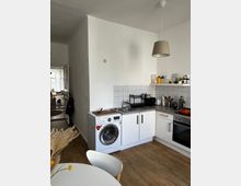 Kleine moderne Küche mit weißen Schränken, grauer Arbeitsplatte und Fliesenspiegel; ausgestattet mit Waschmaschine, Spüle, Herd und Backofen. Im Vordergrund befindet sich ein runder Esstisch und im Hintergrund ein heller Wohnbereich.