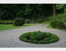 Gepflegter Gartenbereich mit einer kreisförmigen Pflasterung, in deren Mitte ein Beet mit kugelförmig geschnittenen Sträuchern liegt. Im Hintergrund befindet sich eine Wiese mit weiteren Sträuchern und hohen Bäumen, die an einen Wald grenzen.