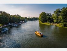 Flusslandschaft mit einem gelben Freizeitboot im Vordergrund, umgeben von dicht bewachsenen Ufern. Links am Ufer liegen mehrere Boote an einer Anlegestelle, im Hintergrund ist ein Turm sichtbar, eingebettet in die grüne Umgebung.