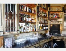 Eine Bar mit einer rustikalen Einrichtung, die Regale voller Spirituosenflaschen, Gläser und Weinflaschen zeigt. Die Theke ist mit gestapelten Trinkgläsern sowie einer Registrierkasse und weiteren Utensilien ausgestattet.