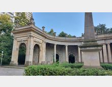 Sandstein-Pavillon mit klassischen Rundbögen und Säulen, verziert mit Reliefs, umgeben von Grünflächen und Bäumen; im Vordergrund steht ein hoher Obelisk. Im Hintergrund sind moderne Gebäude sichtbar.