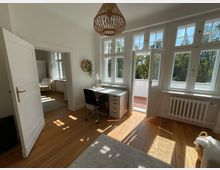 Helles Zimmer mit Holzfußboden, einem Schreibtisch und Stuhl vor einer großen Fensterfront mit Glastür zu einem Balkon. Rechts befindet sich ein Heizkörper, links ist eine offene Tür zu einem angrenzenden Raum sichtbar.