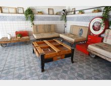 Ein rustikaler Raum mit weiß gekachelten Wänden, die mit blauen Mustern verziert sind. Möbel aus Holzpaletten mit Jutesackbezügen, ein massiver Holztisch in der Mitte und eine rote Waage ergänzen die Einrichtung, während grüne Pflanzen und Dekorationsstücke den Raum schmücken.