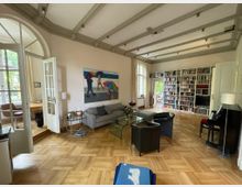 Wohnzimmer mit Holzboden und weißen Wänden, ausgestattet mit einer grauen Couch, schwarzem Klavier, Glastisch, Büchersammlung in hohen weißen Regalen und einem farbenfrohen Gemälde. Große Fenster und Doppeltüren sorgen für viel Tageslicht; rechts daneben ein Durchgang zu einem Arbeitsbereich.