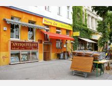 Straßenszene mit einem gelb-orange gestrichenen Gebäude, das ein Antiquitätengeschäft und ein vietnamesisches Restaurant beherbergt; Schilder mit den Aufschriften „Antiquitäten“ und „Huong Que – Vietnamesische Küche“ sind sichtbar. Vor dem Restaurant stehen gestapelte Holztische und -stühle sowie Heizstrahler, und die Fassade ist teilweise mit grünen Kletterpflanzen bedeckt.