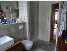 Ein Badezimmer mit grauem Boden und weißen Fliesenwänden. Zu sehen sind ein Waschbecken mit Holzunterschrank, eine Toilette, eine Glasdusche und diverse Pflegeprodukte auf einer Ablage; im Hintergrund führt eine offene Tür in einen weiteren Raum mit Holzboden.