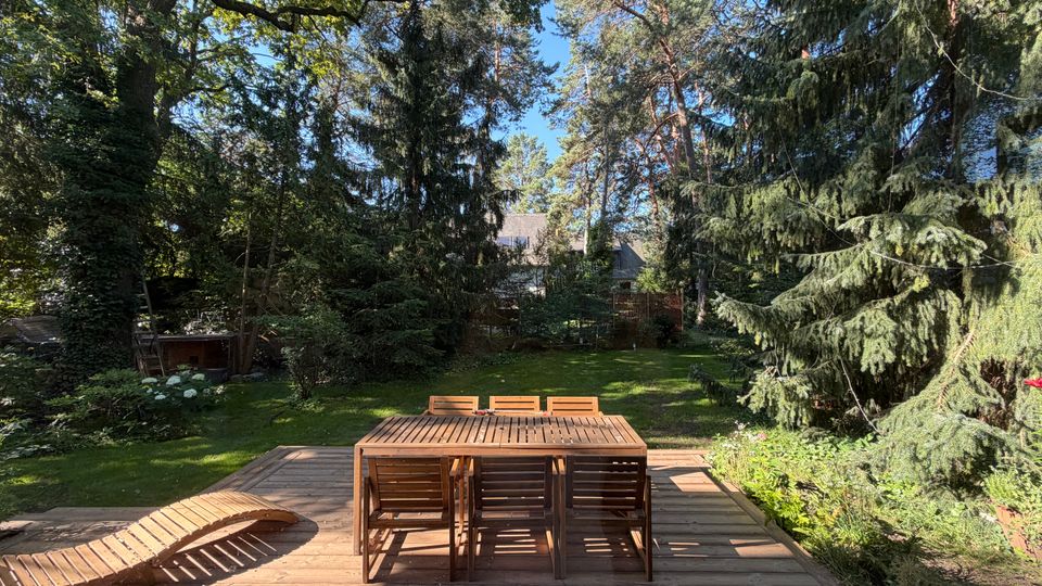 Holzterrasse mit Esstisch, mehreren Stühlen und einer geschwungenen Holzliege, die in einen großen, grünen Garten führt. Der Garten ist von hohen Laub- und Nadelbäumen umgeben, mit Rasenfläche, Sträuchern und einem teilweise sichtbaren Haus im Hintergrund.