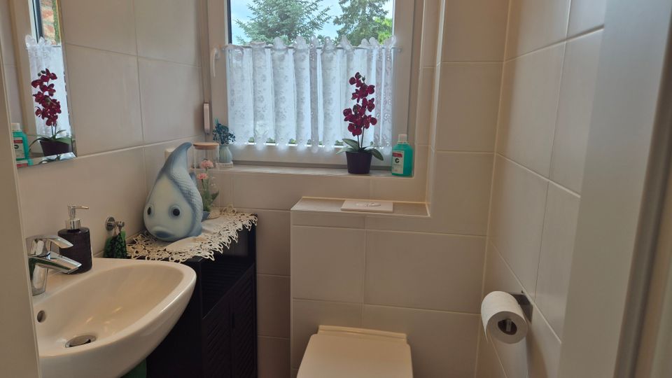 Kleines, hell gefliestes Badezimmer mit Waschbecken links, WC in der Mitte und einem Fenster mit Spitzenvorhang. Auf dem Waschtisch und der Fensterbank stehen Deko und Pflanzen, darunter eine Orchidee; an der rechten Wand ist ein Toilettenpapierhalter montiert.