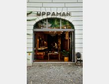 Außenansicht eines modernen Friseursalons mit breitem Eingang und großen Glasfenstern unter einem Bogengiebel, auf dem "UPPAMAN" steht. Im Inneren sichtbar sind Ledersessel, ein Holztisch, Backsteinwände und eine Kronleuchterbeleuchtung.