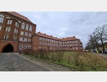 Großes, mehrstöckiges Backsteinschulgebäude mit roten Ziegeldächern und vielen symmetrisch angeordneten Fenstern, links mit einem spitzbogigen Eingang. Davor liegen ein gepflasterter Weg und eine Rasenfläche mit niedrigen Sträuchern; am Rand stehen kahle Bäume und eine Straße.