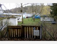 Holztor mit der Hausnummer 31 führt zu einer Wiese mit vereinzelten Bäumen, darauf stehen kleine weiße Gebäude mit flachen Dächern. Im Hintergrund sind ein Fluss mit geparkten Booten und eine bewaldete Uferlandschaft zu sehen.