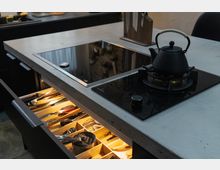 Moderne Küche mit einer Arbeitsplatte aus Beton, die ein Kochfeld mit integriertem Gas- und Induktionsherd enthält. Im Vordergrund befindet sich eine geöffnete Schublade mit beleuchteten, sortierten Küchenutensilien, daneben eine schwarze Teekanne auf dem Gasherd.