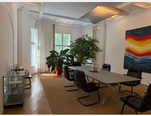 Tagungsraum mit moderner Einrichtung, bestehend aus einem rechteckigen Tisch und schwarzen Stühlen auf einem hellen Teppich. Große Fenster lassen Tageslicht herein, eine grüne Pflanze in einem roten Topf steht in der Ecke, und an der Wand hängt ein farbiges abstraktes Gemälde.