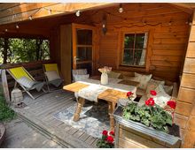 Überdachte Holzterrasse einer Blockhütte mit Holzdecke und Lichterkette, offener Tür und Fenster. Auf der Veranda stehen zwei Liegestühle, eine Eckbank mit Kissen und ein Holztisch mit Tischläufer und Vase; vorn ein Blumenkasten mit roten Geranien.