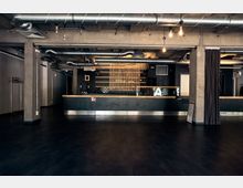 Industrie-inspirierter Innenraum mit dunklem Boden, freiliegenden Rohren an der Decke und einer langen, modernen Bar aus Metall und Holz. Im Hintergrund sind offene Regale, ein Sichtschutz aus Holzlatten und schlichte Wände erkennbar.