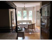 Wohnzimmer mit einem Essbereich im klassischen Stil, bestehend aus einem runden Tisch mit weißer Tischdecke, Stühlen und einer gepolsterten Sitzbank. Im Hintergrund befindet sich eine große Fensterfront, flankiert von weißen, eingebauten Vitrinenschränken, und der Boden ist aus Holz.