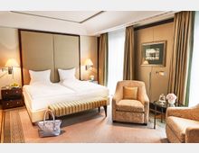 Elegantes Hotelzimmer mit einem Doppelbett, gepolstertem Kopfteil und beidseitigen Nachttischen mit Tischlampen. Der Raum ist mit einem Sessel, einem Beistelltisch mit Blumen und einem großen Fenster mit Vorhängen ausgestattet; beige- und goldfarbene Töne dominieren das Interieur.